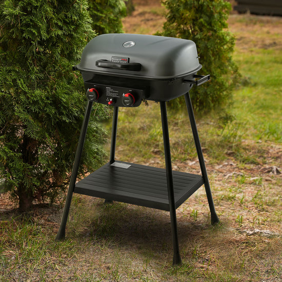 A black Royal Gourmet portable gas grill with a lid, two legs, and a lower shelf, set outdoors on grass next to green shrubs. The grill has red control knobs on the front.