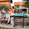 A lifestyle scene at a poolside backyard where a group of four friends are gathered around a black Royal Gourmet gas grill cooking food. The setting features a stone patio, a swimming pool, and a house in the background.