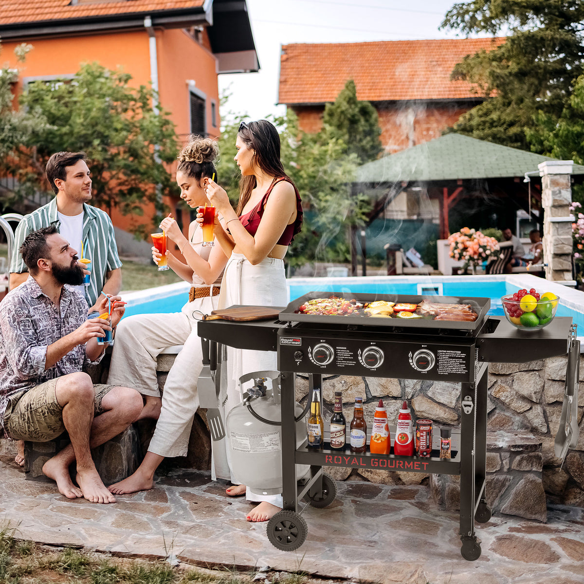 A lifestyle scene at a poolside backyard where a group of four friends are gathered around a black Royal Gourmet gas grill cooking food. The setting features a stone patio, a swimming pool, and a house in the background.