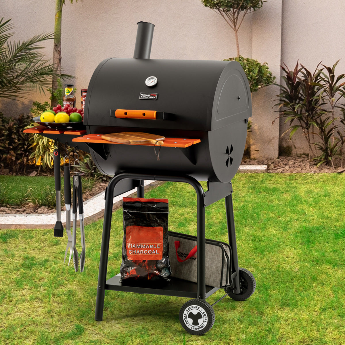 A black Royal Gourmet charcoal grill with a side table, hanging utensils, and a bag of Hammerbell Charcoal on a green lawn in front of a beige stucco wall.