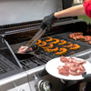 A close-up lifestyle shot of a two-burner Royal Gourmet gas grill in use. A person wearing a red shirt and a black glove uses metal tongs to place a piece of meat into a cast iron skillet on the left grill grate.