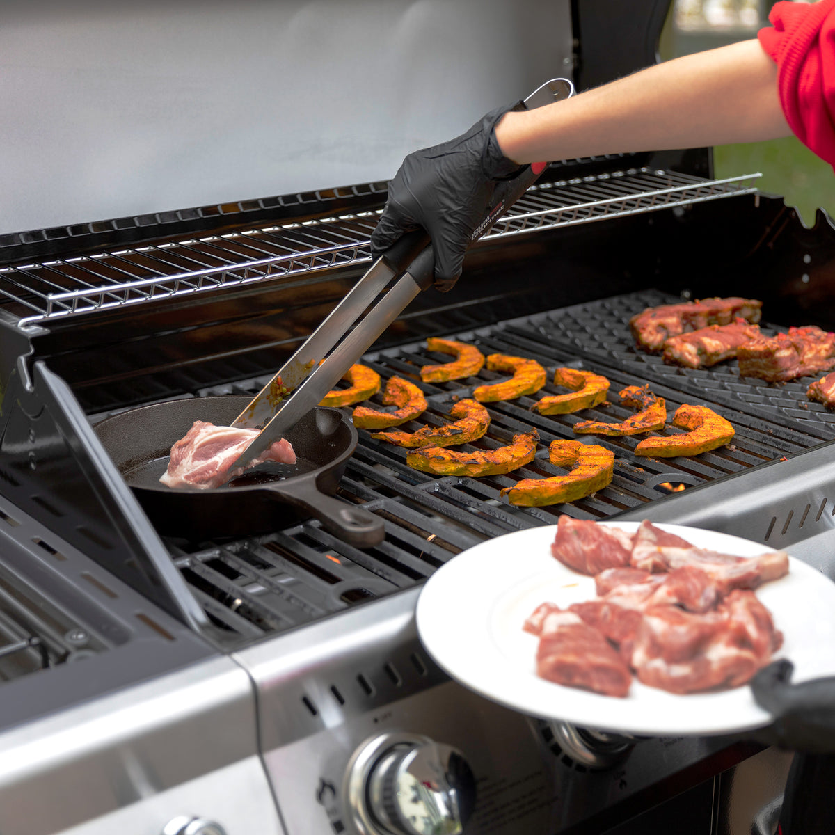 A close-up lifestyle shot of a two-burner Royal Gourmet gas grill in use. A person wearing a red shirt and a black glove uses metal tongs to place a piece of meat into a cast iron skillet on the left grill grate.