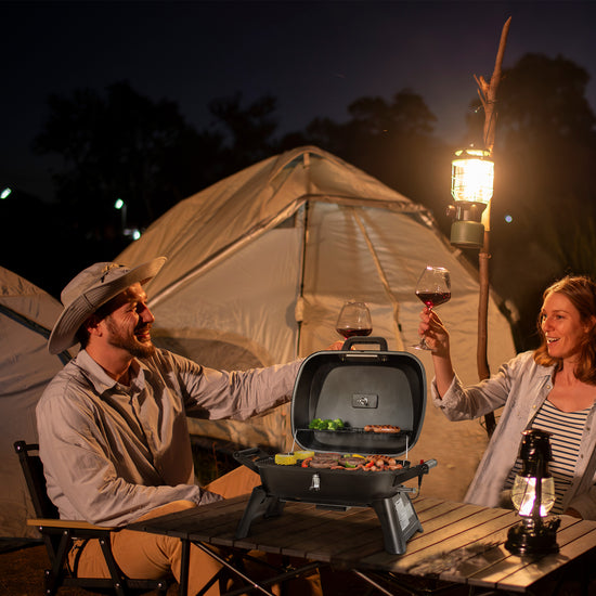A man and woman toast at a campsite nightstand with a portable Royal Gourmet charcoal grill, wine glasses, and a lantern, with a large canvas tent illuminated by warm lights in the background.