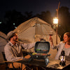 A man and woman toast at a campsite nightstand with a portable Royal Gourmet charcoal grill, wine glasses, and a lantern, with a large canvas tent illuminated by warm lights in the background.