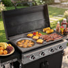 A black and gray ProGrill Royal Gourmet gas grill with the lid open, cooking burgers, steak, corn, peppers, and fruit outdoors on a grassy lawn. Condiment bottles are lined up on the right side.