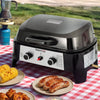 A black and silver Royal Gourmet outdoor countertop grill sits on a red-and-white checkered picnic blanket outdoors, with plates of corn, sausages, and chicken beside it. A blue gas canister and metal pot are also visible in the scene.