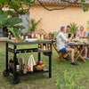 Outdoor setting with a barbecue cart, people sitting at a table, and a yellow building in the background.