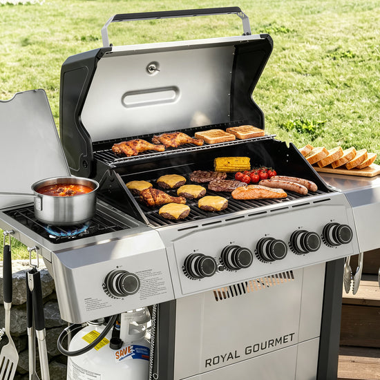 A Royal Gourmet gas grill set outdoors on a wooden deck with a grassy lawn in the background. The grill is open, showing various foods cooking on the grates and side shelf, including steak, sausages, corn, tomatoes, and a pot of soup.