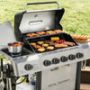 A Royal Gourmet gas grill set outdoors on a wooden deck with a grassy lawn in the background. The grill is open, showing various foods cooking on the grates and side shelf, including steak, sausages, corn, tomatoes, and a pot of soup.