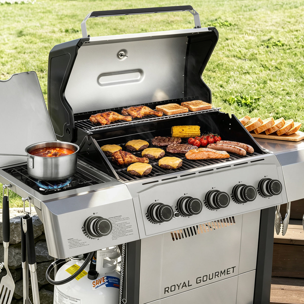 A Royal Gourmet gas grill set outdoors on a wooden deck with a grassy lawn in the background. The grill is open, showing various foods cooking on the grates and side shelf, including steak, sausages, corn, tomatoes, and a pot of soup.