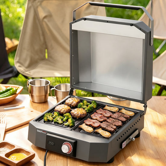 A compact Royal Gourmet electric grill with a hinged lid open, cooking steak, broccoli, mushrooms, and chicken on a wooden table outdoors. Stainless steel cups, a cutting board, and camping chairs are visible in the background.