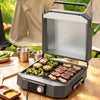 A compact Royal Gourmet electric grill with a hinged lid open, cooking steak, broccoli, mushrooms, and chicken on a wooden table outdoors. Stainless steel cups, a cutting board, and camping chairs are visible in the background.