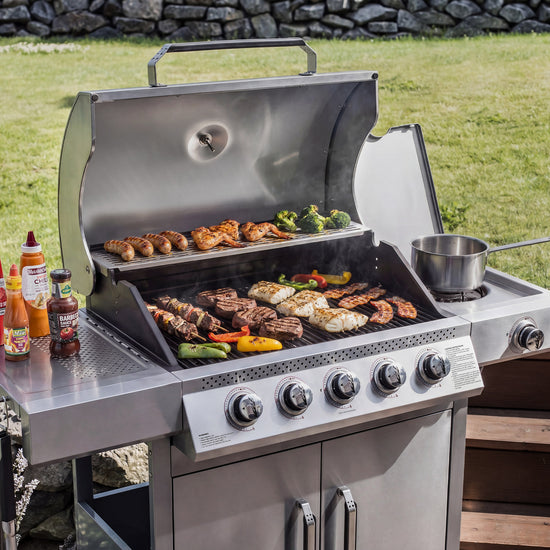 A stainless steel Royal Gourmet gas grill with multiple food items cooking on the grates and a side shelf with condiment bottles, set outdoors on a wooden deck with grass and a stone wall in the background.