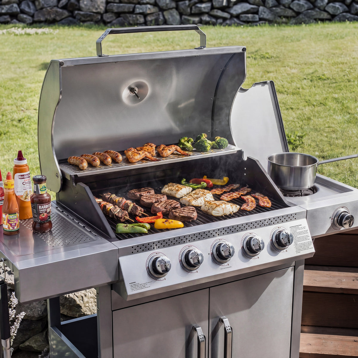 A stainless steel Royal Gourmet gas grill with multiple food items cooking on the grates and a side shelf with condiment bottles, set outdoors on a wooden deck with grass and a stone wall in the background.