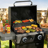 A black outdoor Royal Gourmet gas grill with a lid handle and side table, shown open and cooking various foods like burgers, steak, corn, and vegetables. Condiment bottles and sliced bread are placed on the side table, with a stone wall and grassy background.