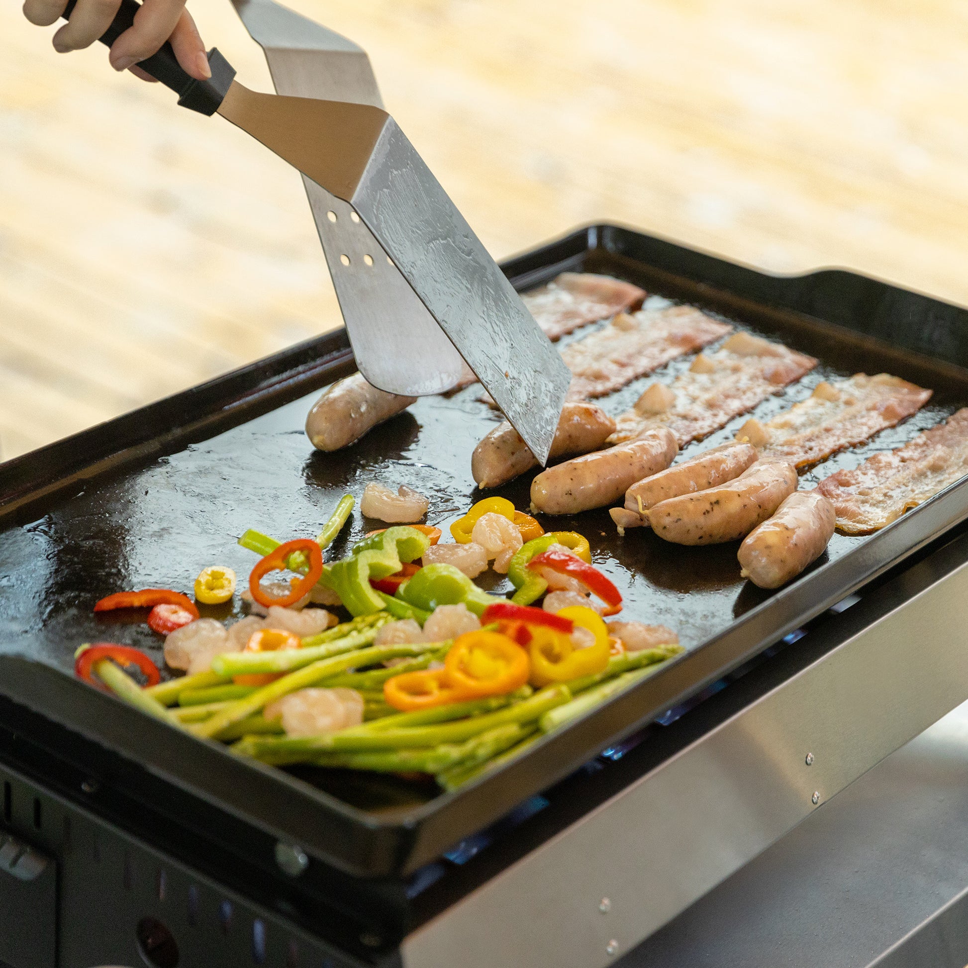Person grilling sausages and vegetables on a Royal Gourmet portable grill with a spatula.