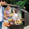 A black Royal Gourmet outdoor gas grill is open on a patio with three people grilling food, including burgers, steak, corn, and peppers, with smoke rising and a garden background.