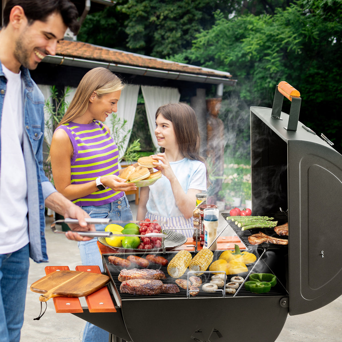 A black Royal Gourmet outdoor gas grill is open on a patio with three people grilling food, including burgers, steak, corn, and peppers, with smoke rising and a garden background.