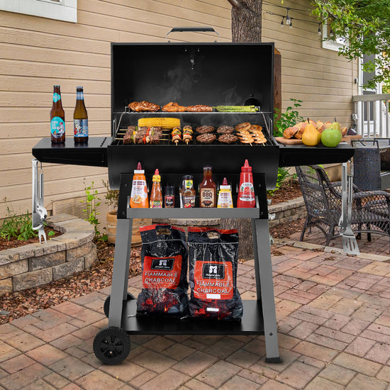 A black Royal Gourmet charcoal grill with a side shelf and lower storage shelf, shown open on a brick patio in front of a house. The grill is loaded with food, with bottles of sauce and condiments on the side shelf and two bags of HZ Premium Charcoal in the lower compartment.