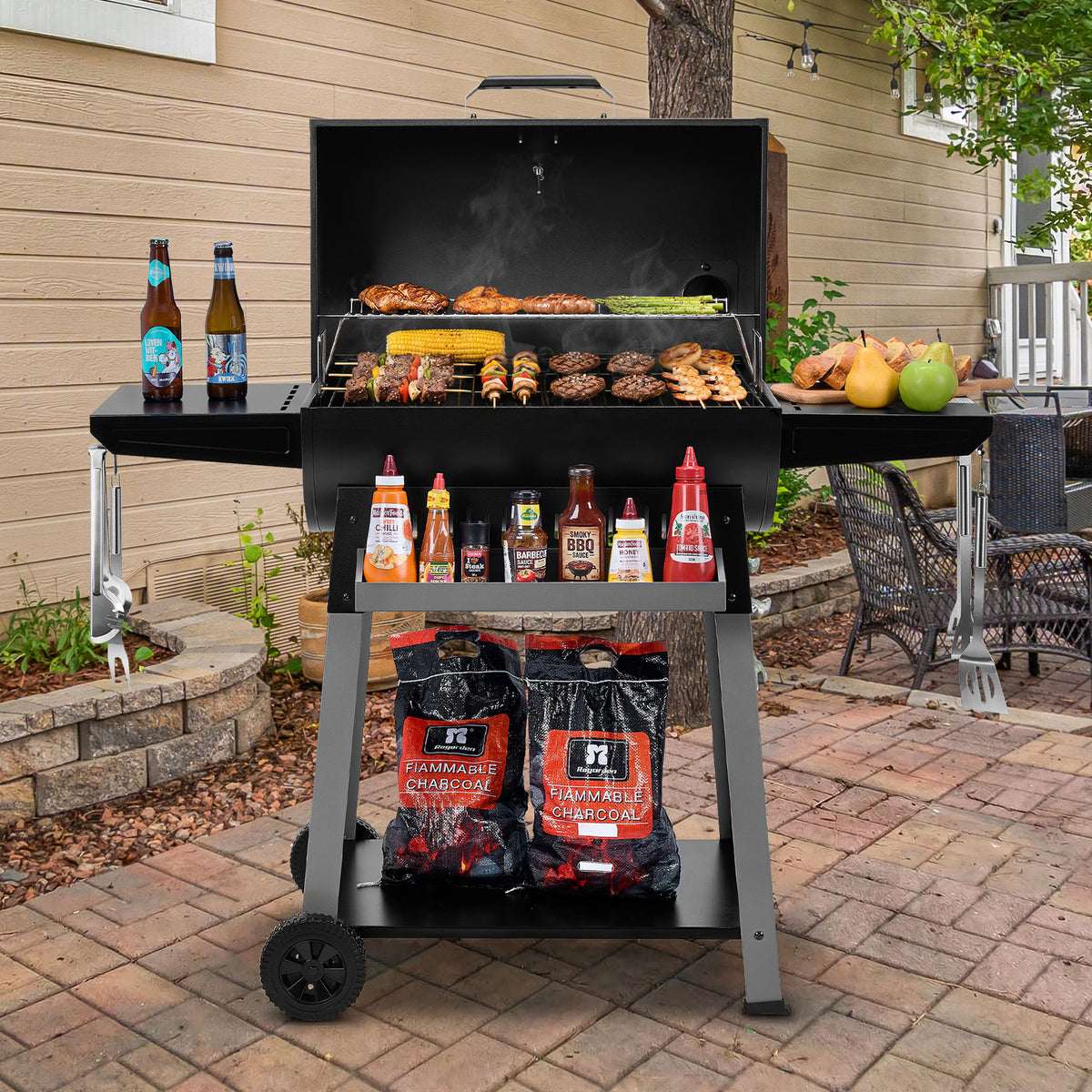 A black Royal Gourmet charcoal grill with a side shelf and lower storage shelf, shown open on a brick patio in front of a house. The grill is loaded with food, with bottles of sauce and condiments on the side shelf and two bags of HZ Premium Charcoal in the lower compartment.