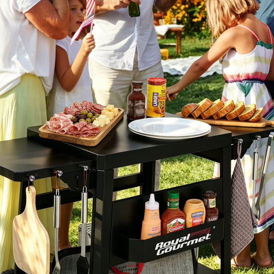 A black Royal Gourmet outdoor kitchen cart with a side table top, hanging utensils, and a lower storage basket, shown in use outdoors with people serving food around it.