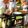 A black Royal Gourmet outdoor kitchen cart with a side table top, hanging utensils, and a lower storage basket, shown in use outdoors with people serving food around it.