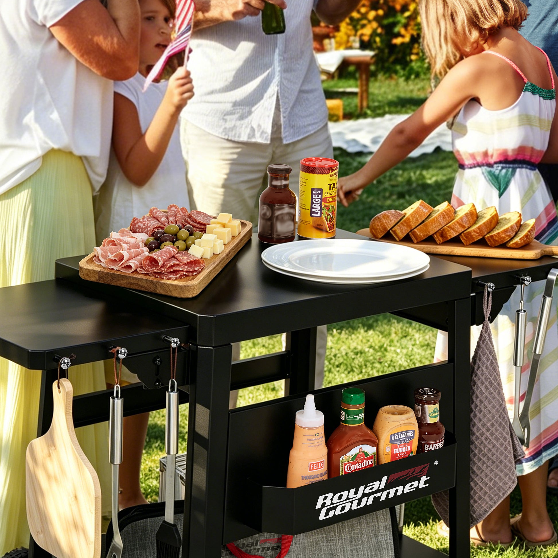 A black Royal Gourmet outdoor kitchen cart with a side table top, hanging utensils, and a lower storage basket, shown in use outdoors with people serving food around it.