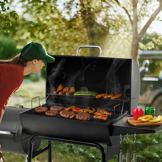 A black outdoor Royal Gourmet  charcoal grill is shown with meat and vegetables cooking on the grates, with smoke rising. A person wearing a green cap and red shirt leans in from the left, and a garden background with trees and a vehicle is visible.