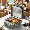 A portable electric Royal Gourmet  grill with a lid is shown open on a patio table, cooking steak, sausage, asparagus, pineapple, and skewers. People are seated at a dining table in the background, and ice-cold beer bottles sit in a bucket in the foreground.