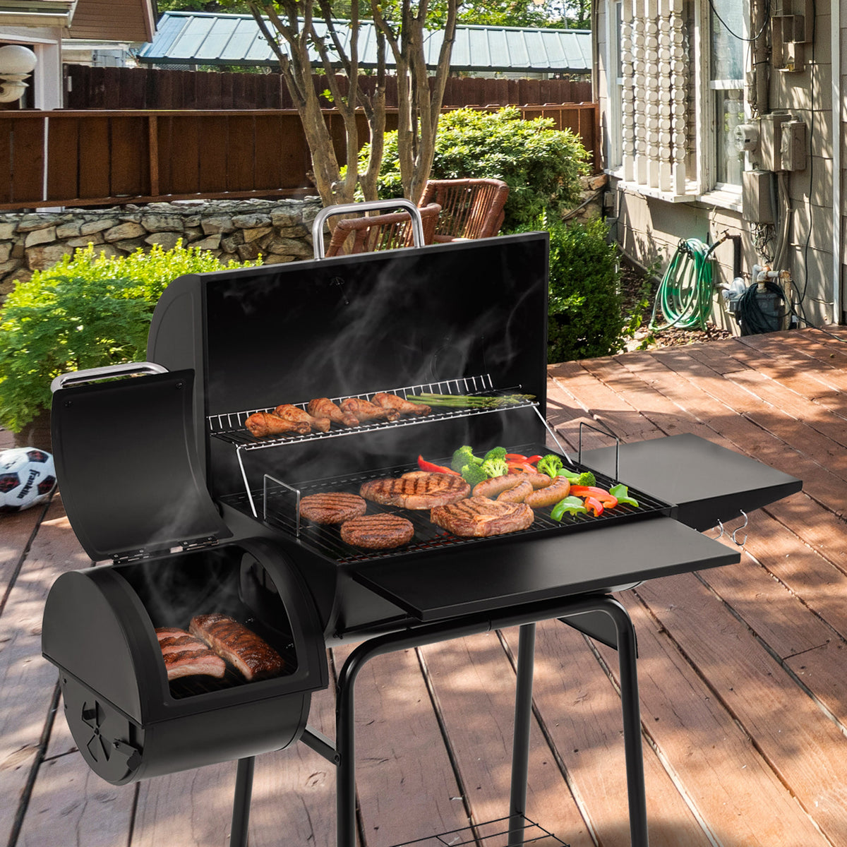A black Royal Gourmet  outdoor pellet grill with a lid, side table, and lower fire pot, shown cooking sausages, burgers, and vegetables on a wooden deck in a backyard setting.