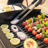 A close-up of a black Royal Gourmet   grill with skewers of chicken, red tomatoes, yellow and green bell peppers, sliced red onion, lemon halves, and a halved avocado. Metal tongs with black handles are lifting a slice of red onion over the grates.