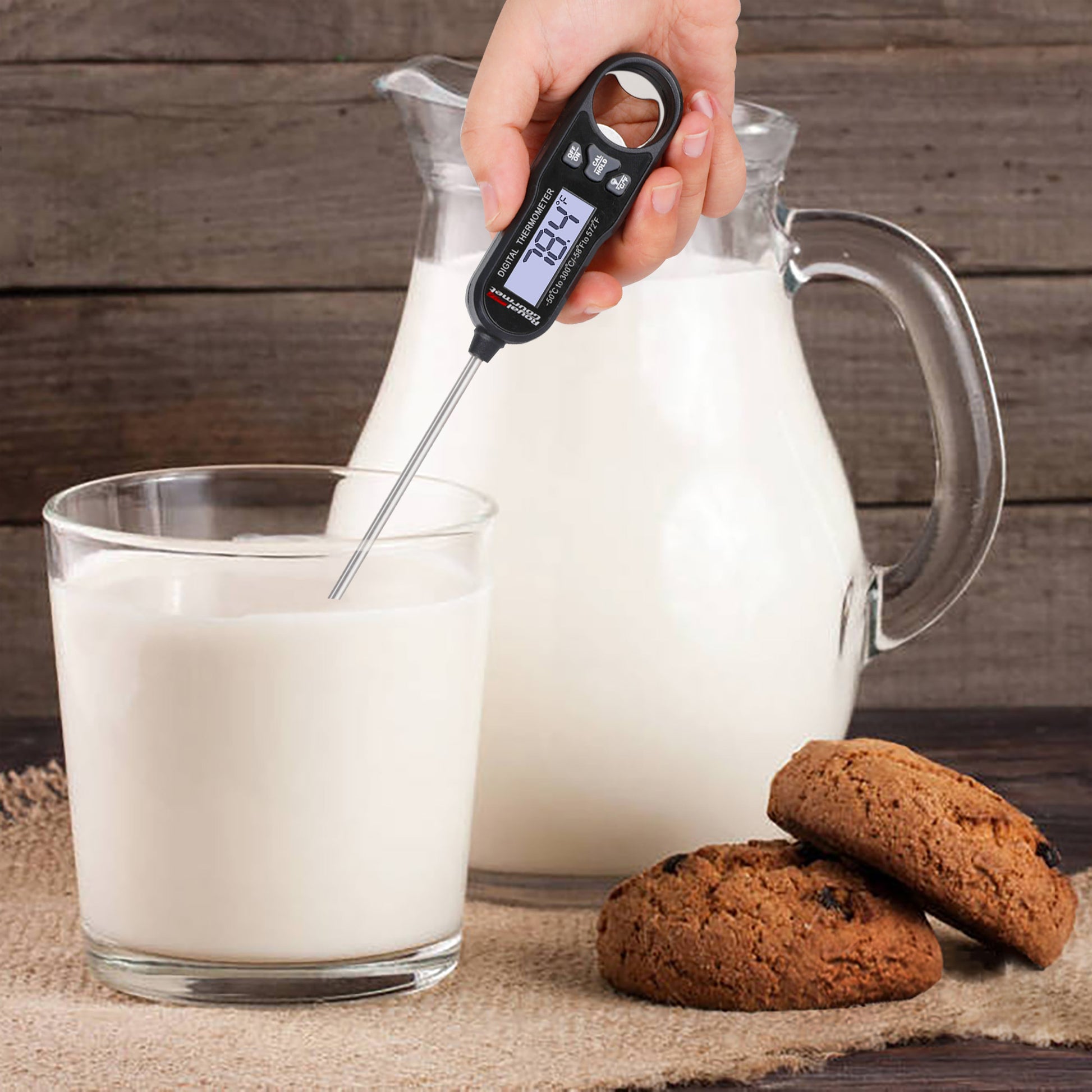 Glass of milk with a digital Royal Gourmet thermometer, pitcher of milk, and cookies on a wooden surface.