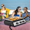 People enjoying a picnic with a Royal Gourmet portable grill on a checkered blanket by the beach.