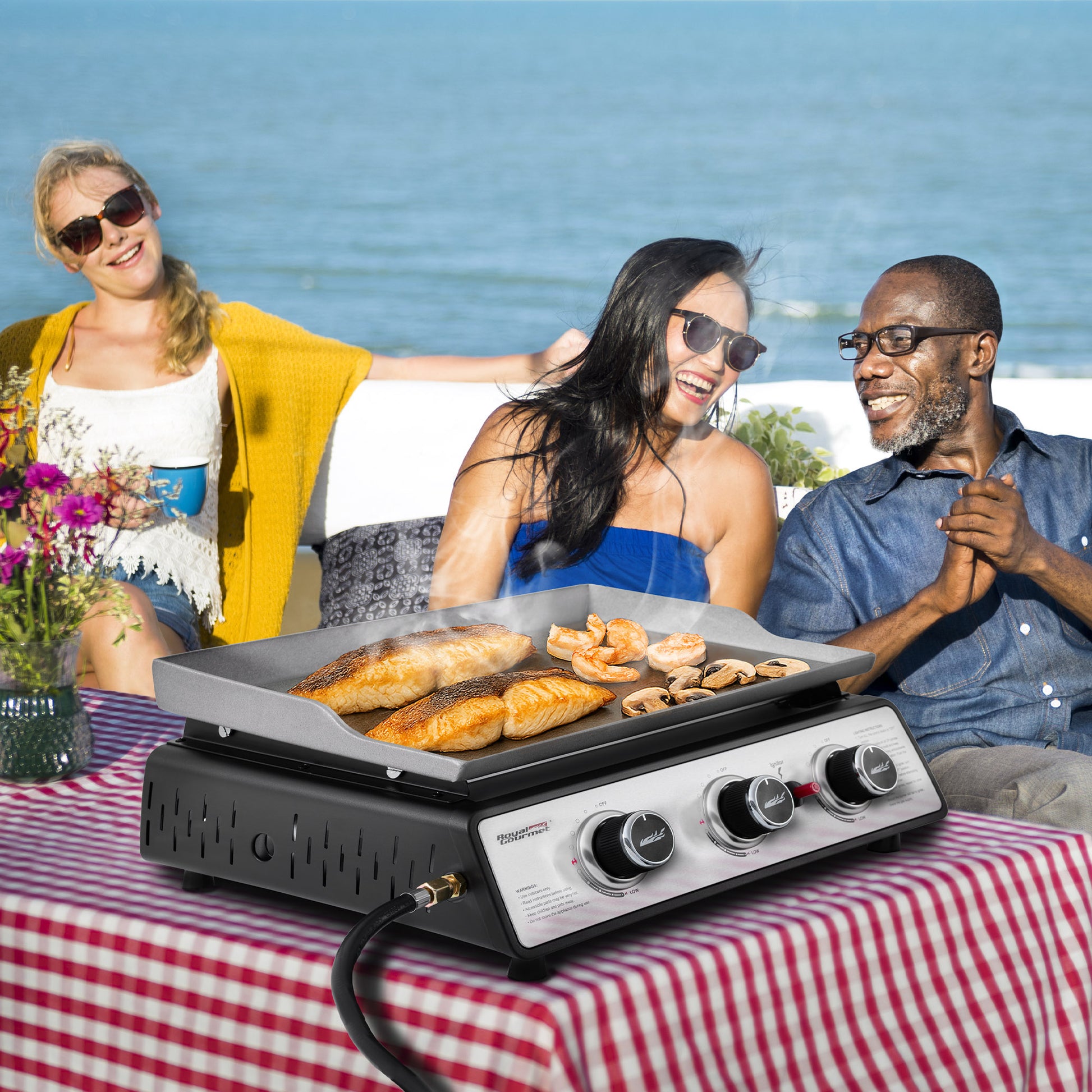 People enjoying a picnic with a Royal Gourmet portable grill on a checkered blanket by the beach.