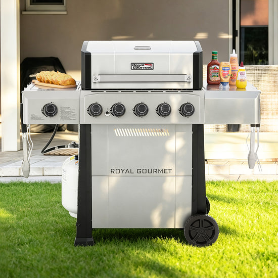 A Royal Gourmet gas grill with side shelves, set on a green lawn in front of a patio. The grill is white and black with five control knobs on the front, a propane tank connected underneath, and food and condiments on its side tables.