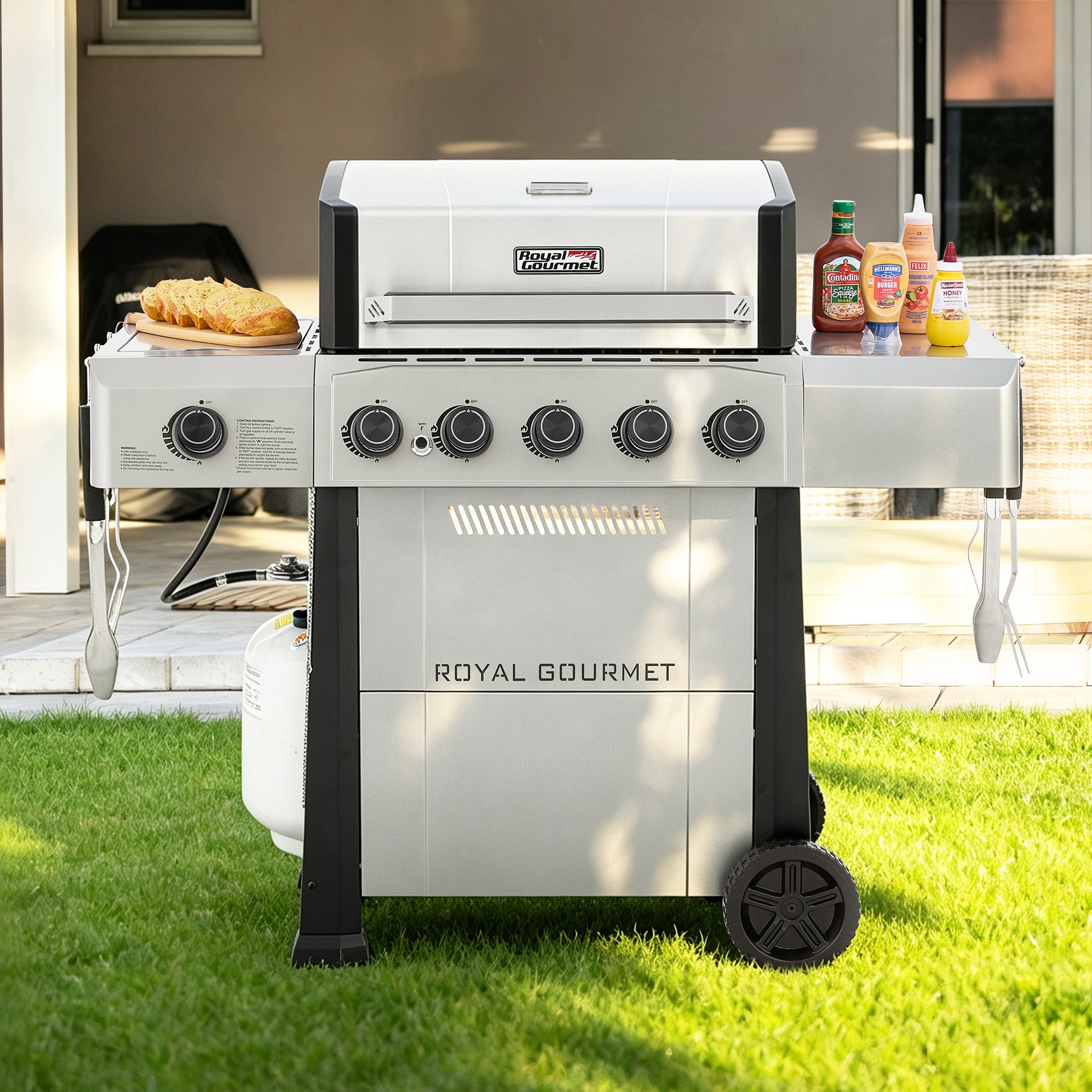 A Royal Gourmet gas grill with side shelves, set on a green lawn in front of a patio. The grill is white and black with five control knobs on the front, a propane tank connected underneath, and food and condiments on its side tables.