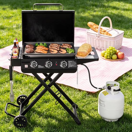 A black Royal Gourmet portable gas grill with a side table is set up on a green grass lawn next to a pink picnic blanket, propane tank, basket, and food items.