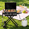 A black Royal Gourmet portable gas grill with a side table is set up on a green grass lawn next to a pink picnic blanket, propane tank, basket, and food items.