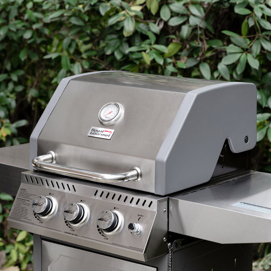 A stainless steel Royal Gourmet gas grill with a gray lid and silver control knobs, photographed outdoors in front of green foliage. The grill has a visible Miller Heat Signature logo on the front.