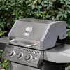 A stainless steel Royal Gourmet gas grill with a gray lid and silver control knobs, photographed outdoors in front of green foliage. The grill has a visible Miller Heat Signature logo on the front.