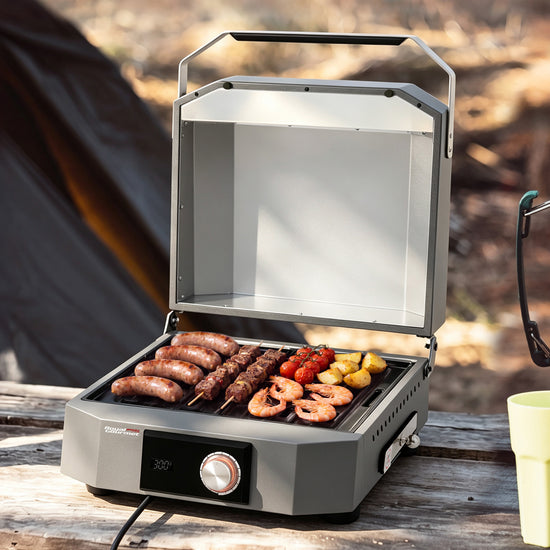 A compact Royal Gourmet electric grill with its lid open, cooking sausages, skewers, shrimp, and vegetables on a wooden table outdoors. The grill is gray with a black control panel and a silver dial.