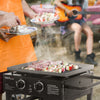 A black outdoor flat-top grill with two knobs, cooking meat and vegetable skewers on a patio. A person in an orange apron is holding plates of food, and another person is seated in the background playing an orange guitar.