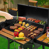 Person grilling various meats, vegetables, and skewers on a portable Royal Gourmet  grill with a wooden bench.