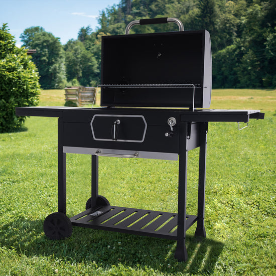 A black Royal Gourmet outdoor charcoal grill with a side table, lower shelf, and wheels, shown on a green grass lawn with trees in the background.