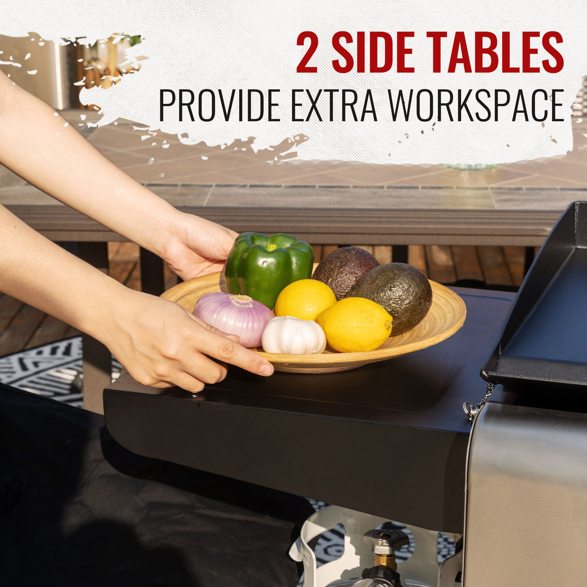 Outdoor lifestyle shot of a person placing a wooden bowl of fruits and vegetables onto a black side table of a grill. Text overlay at the top reads '2 SIDE TABLES PROVIDE EXTRA WORKSPACE'.