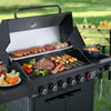 A black Royal Gourmet outdoor gas grill with multiple cooking items on and around it, photographed in a grassy backyard setting. The grill is open, showing sausages on the upper rack, a steak and skewers on the grates, mushrooms, peppers, broccoli, shrimp, and a pot on the side burner.