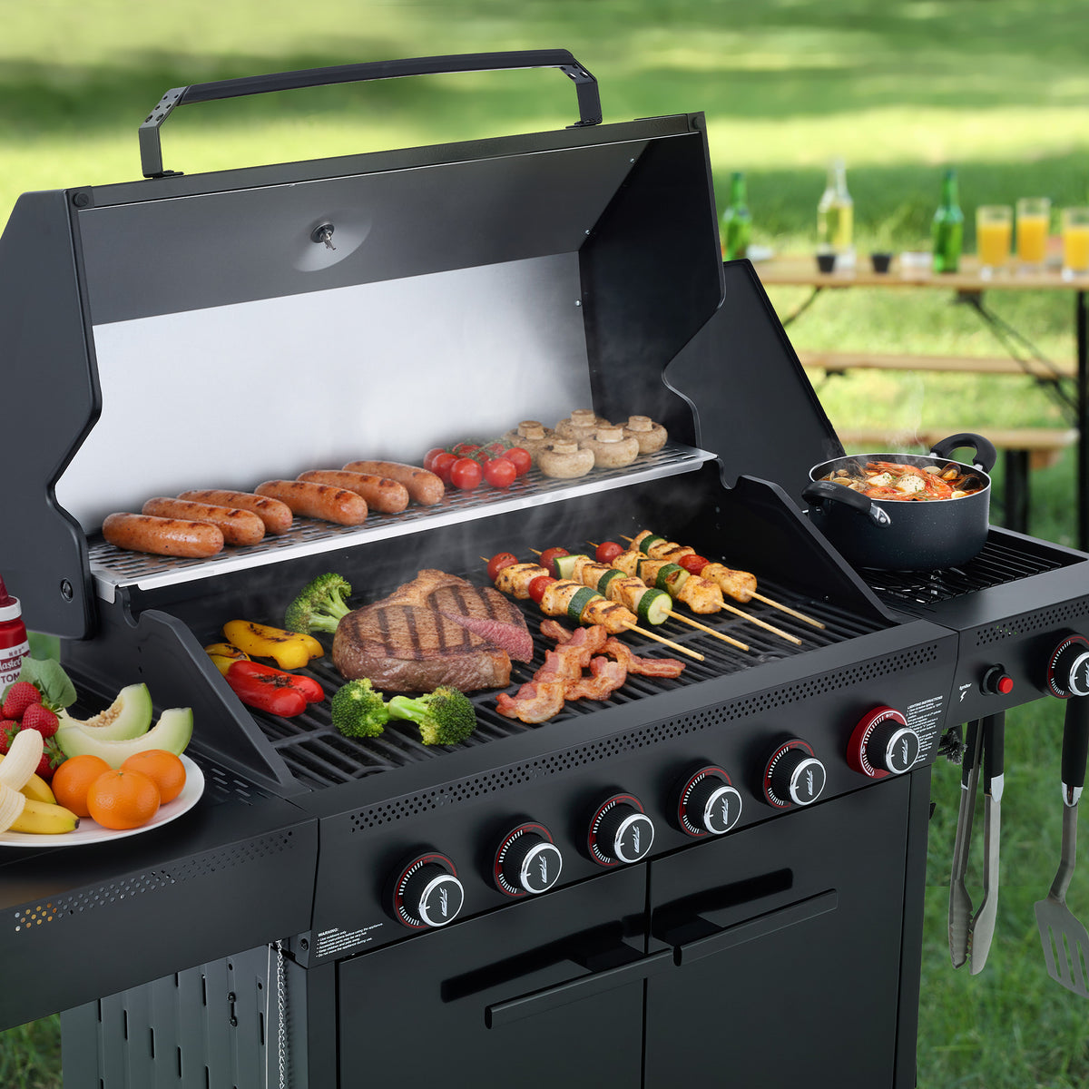 A black Royal Gourmet outdoor gas grill with multiple cooking items on and around it, photographed in a grassy backyard setting. The grill is open, showing sausages on the upper rack, a steak and skewers on the grates, mushrooms, peppers, broccoli, shrimp, and a pot on the side burner.