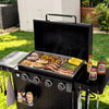 A black outdoor Royal Gourmet gas grill with the lid open, shown on a grassy lawn with food cooking on the griddle and side shelves. Condiment bottles are placed on the side table, and a white shed and garden plants are visible in the background.