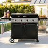 A black and silver outdoor Royal Gourmet gas grill with side tables and accessories, set on a patio in a backyard with a pool and greenery in the background.