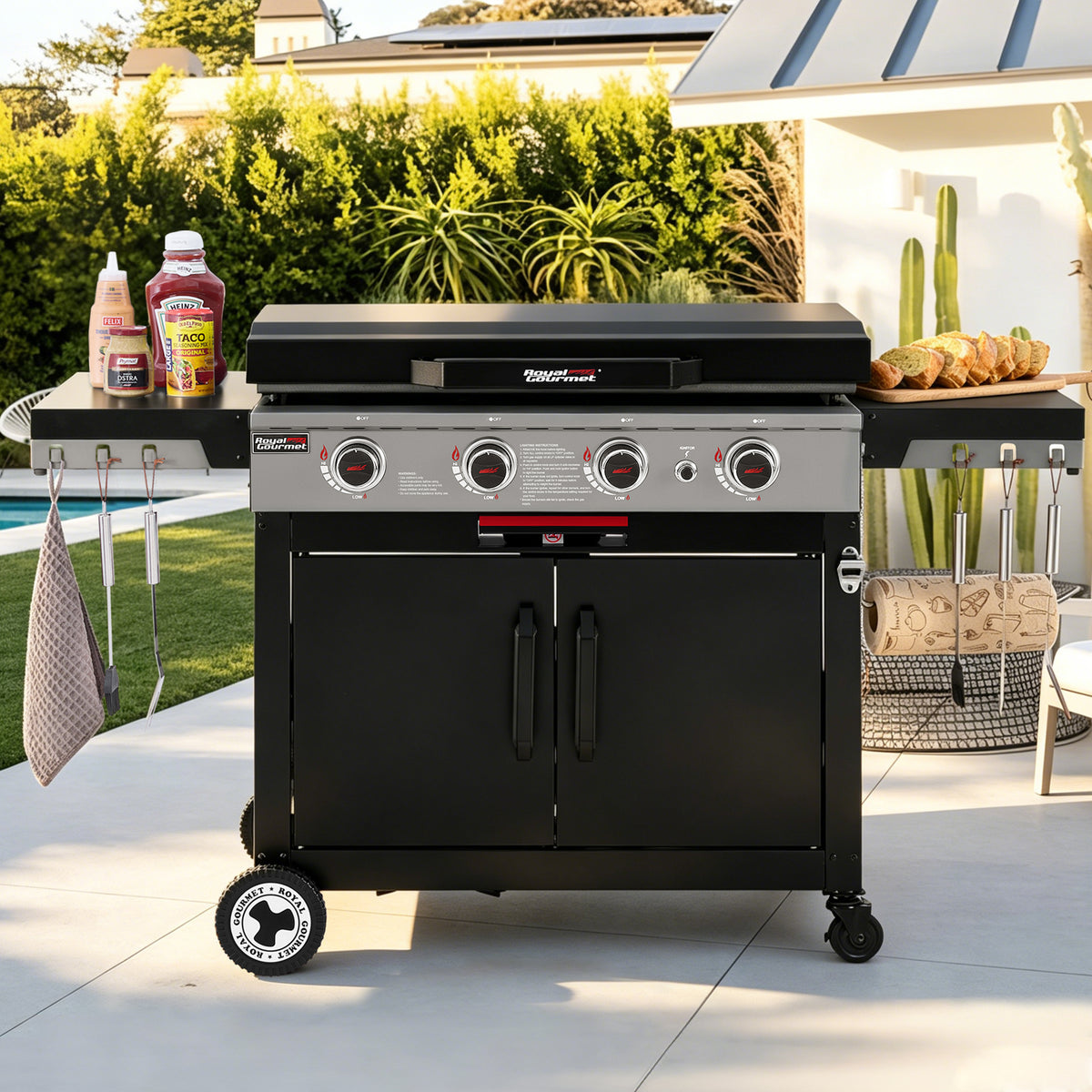 A black and silver outdoor Royal Gourmet gas grill with side tables and accessories, set on a patio in a backyard with a pool and greenery in the background.
